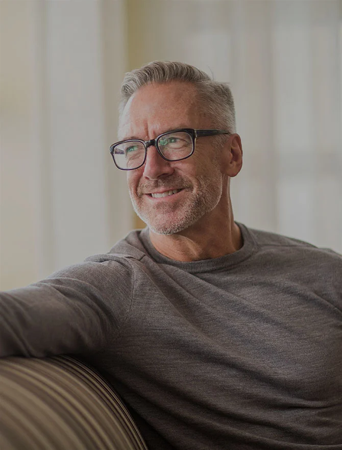 Close-up of a middle-aged white man sitting on a couch with one arm extended on the backrest. He has white hair, light facial hair, is wearing glasses, and a grey long-sleeve shirt. - Procedures in Webster, TX