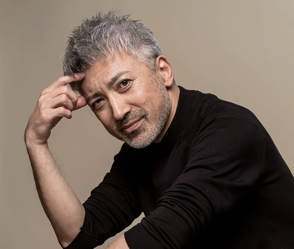 A headshot of an older man resting his head on one hand, with his other hand resting on his lap as he slightly leans forward. He wears a black long-sleeve mockneck shirt and has short grey hair, paired with a buzzed white beard and mustache combo. The light brown blurred background creates a warm, soft ambiance, highlighting his thoughtful expression. - Corporeal Shockwave Lithontripsy Treatment in Webster, TX
