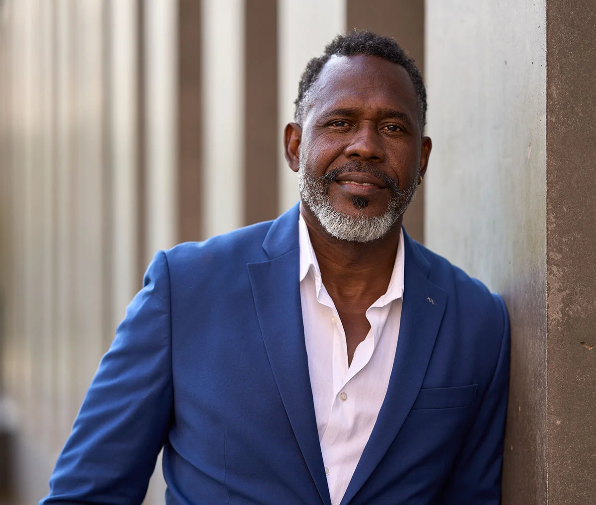 A middle-aged Black man leans casually against a concrete pillar, with a slight smile on his face. He’s dressed in a blue suit over a white button-down shirt, which is unbuttoned at the top for a relaxed look. His black hair and beard are accented with a touch of grey at the tip of his beard, adding a distinguished flair. The background is slightly blurred, revealing a series of concrete columns that create a sense of depth in the scene. - Greenlight Laser Therapy Treatment in Webster, TX