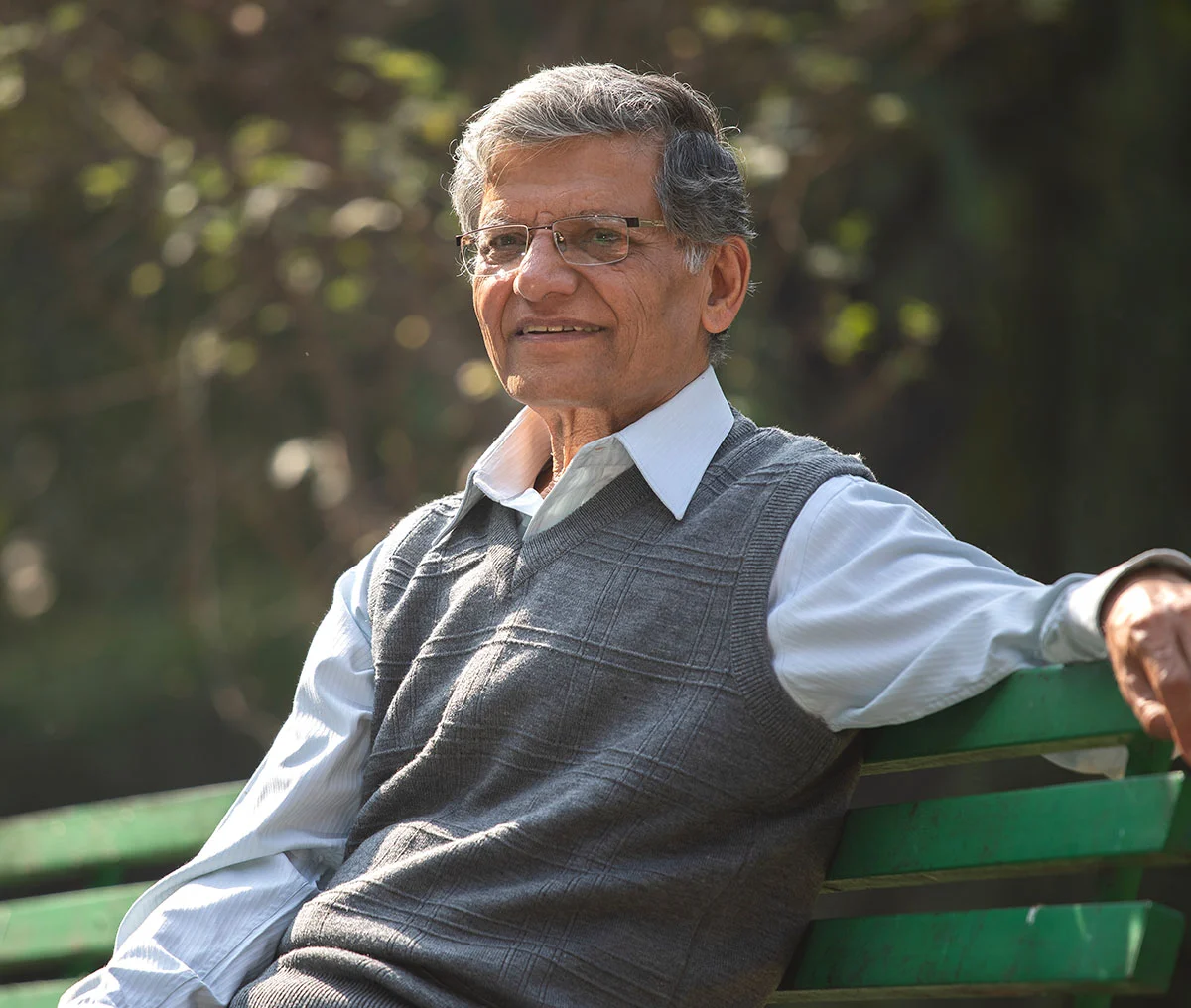 An older male sitting on a park bench, wearing a grey sweater vest over a white collared long-sleeve shirt. He has grey hair and glasses, with one arm resting over the back of the bench. - Prostate Cancer Mention Fusion Biopsy Prostate Biopsy