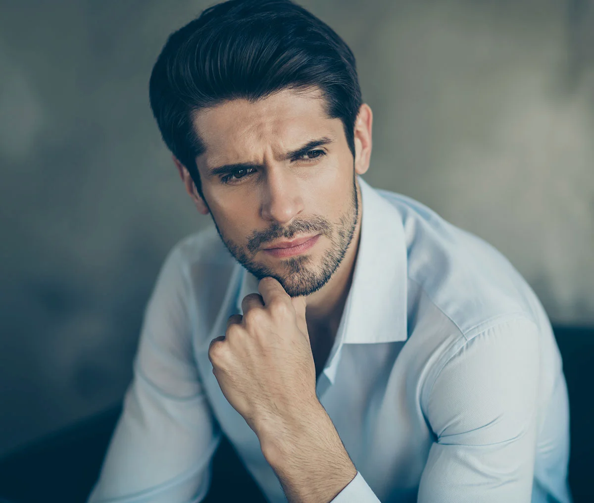 Close-up of a young man with a concerned expression, resting one hand on his chin. He has black hair, a thin beard, and is wearing a white long-sleeve button-down shirt. - Testosterone Replacement Therapy