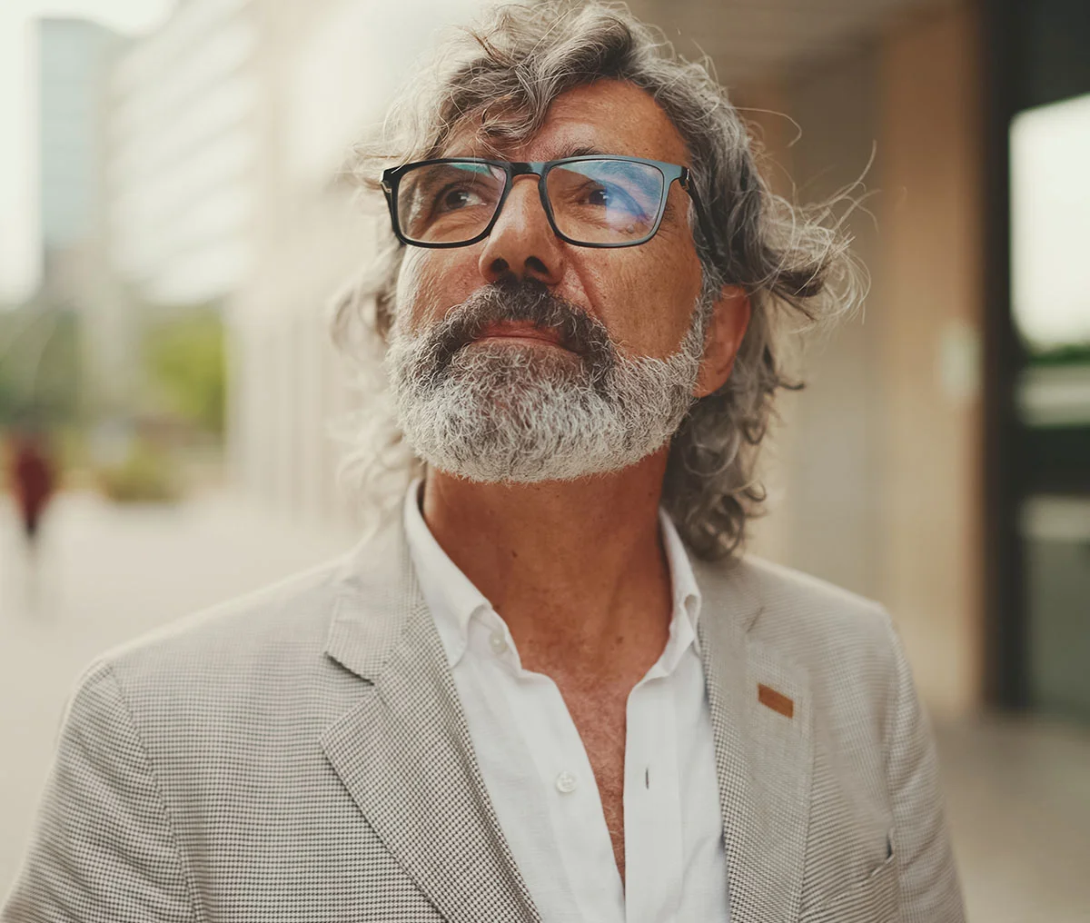 Close up of a man with glasses and a gray beard looks upward while standing outside a building. He wears a light-colored blazer and white shirt. - Home, Webster, TX