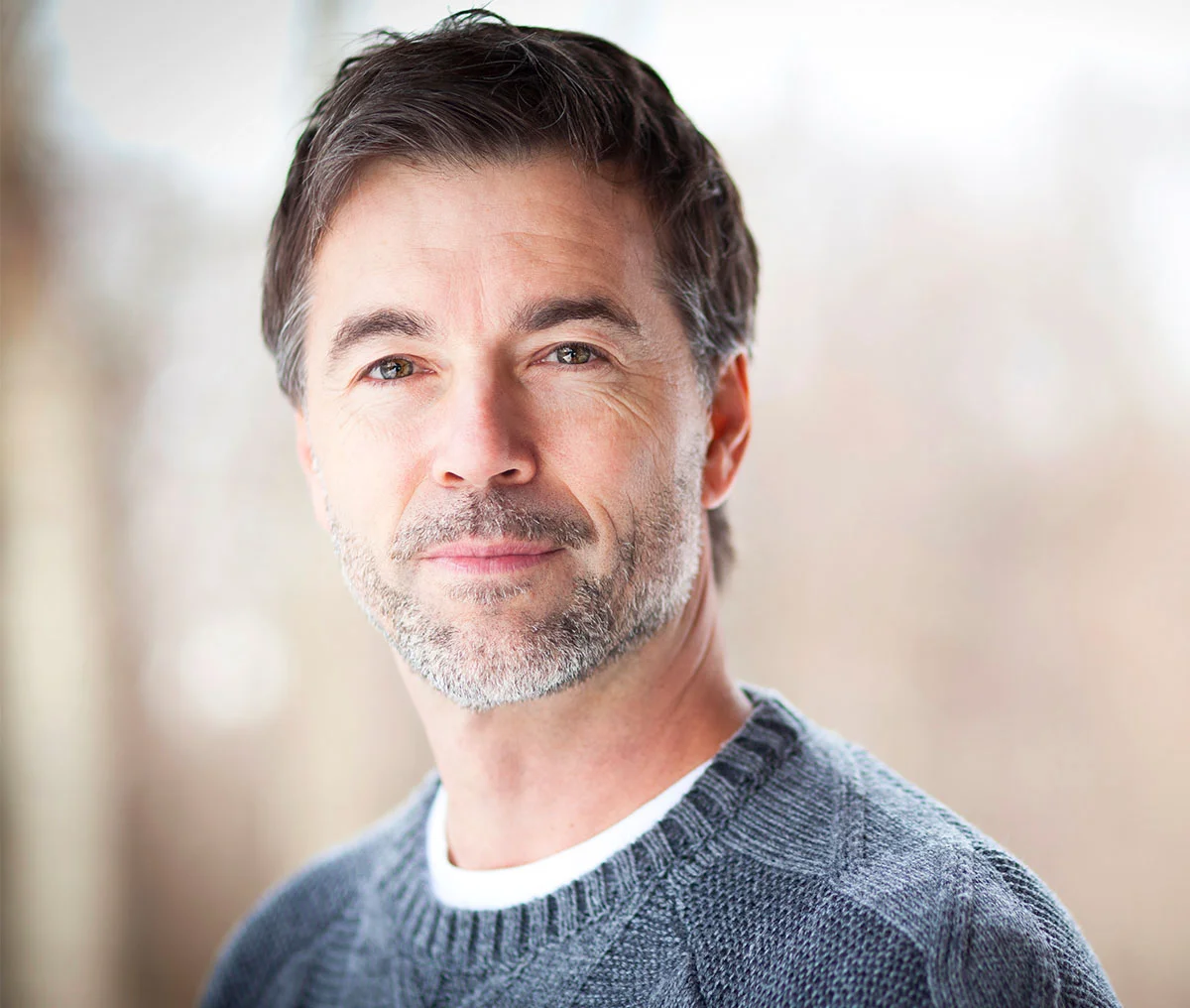 A middle-aged man with light brown hair and a scruffy beard and mustache, flecked with brown and white, looks slightly smiling at the camera. He’s wearing a greyish-blue sweater over a white undershirt, presenting a relaxed and approachable look. The background is softly blurred, keeping the focus on his gentle expression and warm gaze. - Bladder Cancer Treatment in Webster, TX