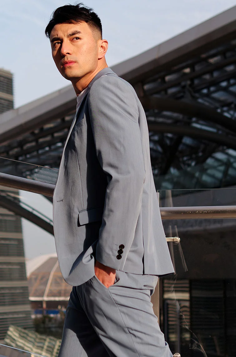 A young man dressed in a grey suit with black buttons and a white undershirt walks through an industrial-style setting, surrounded by raw, architectural buildings. He looks behind and above the camera, his profile captured as the shot faces the side of his body. His hand, visible in the photo, is casually tucked in his pocket. He has black hair that fades, completing a modern, stylish look in the urban environment. - Conditions Treatment in Webster, TX