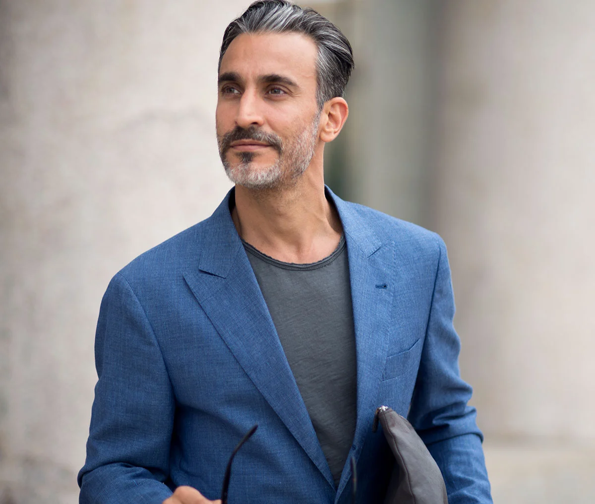 A middle-aged man dressed in a blue suit with a dark grey undershirt stands facing the camera, but with his body angled slightly to the side. He holds sunglasses in one hand and a small black bag under his other arm. His slight grey beard and brown mustache, with touches of grey highlights in his hair, give him a distinguished look. He sports a subtle smile, adding a relaxed yet polished vibe to the scene. - Erectile Dysfunction Treatment in Webster, TX