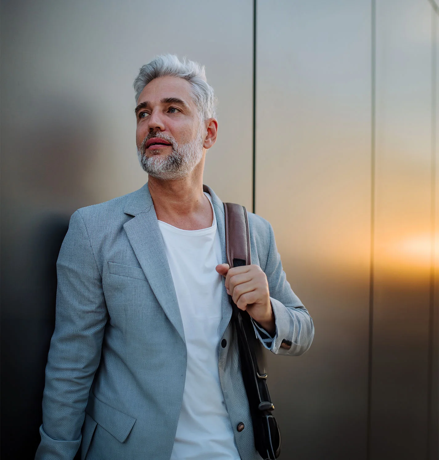 A middle-aged man with a silver beard and hair featuring subtle brown lowlights leans casually against a sleek steel wall, his mirrored reflection softly illuminated by the warm glow of sunset. He carries a men's shoulder bag on his left shoulder, his hand resting on the midpoint of the strap. Dressed in a tailored grey blazer over a clean white T-shirt, he exudes an effortless, polished style in the ambient lighting. - Home. Webster, TX