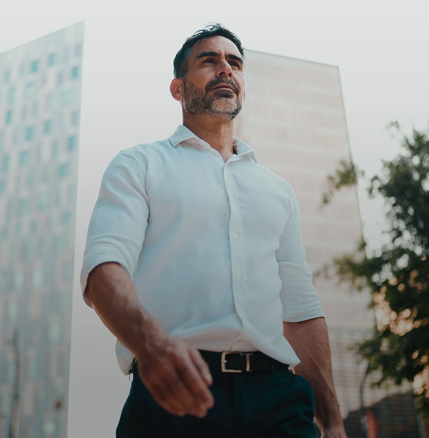 A man in a white shirt and black pants walks outdoors near tall buildings, with a tree in the background. - Mens Health in Webster, TX