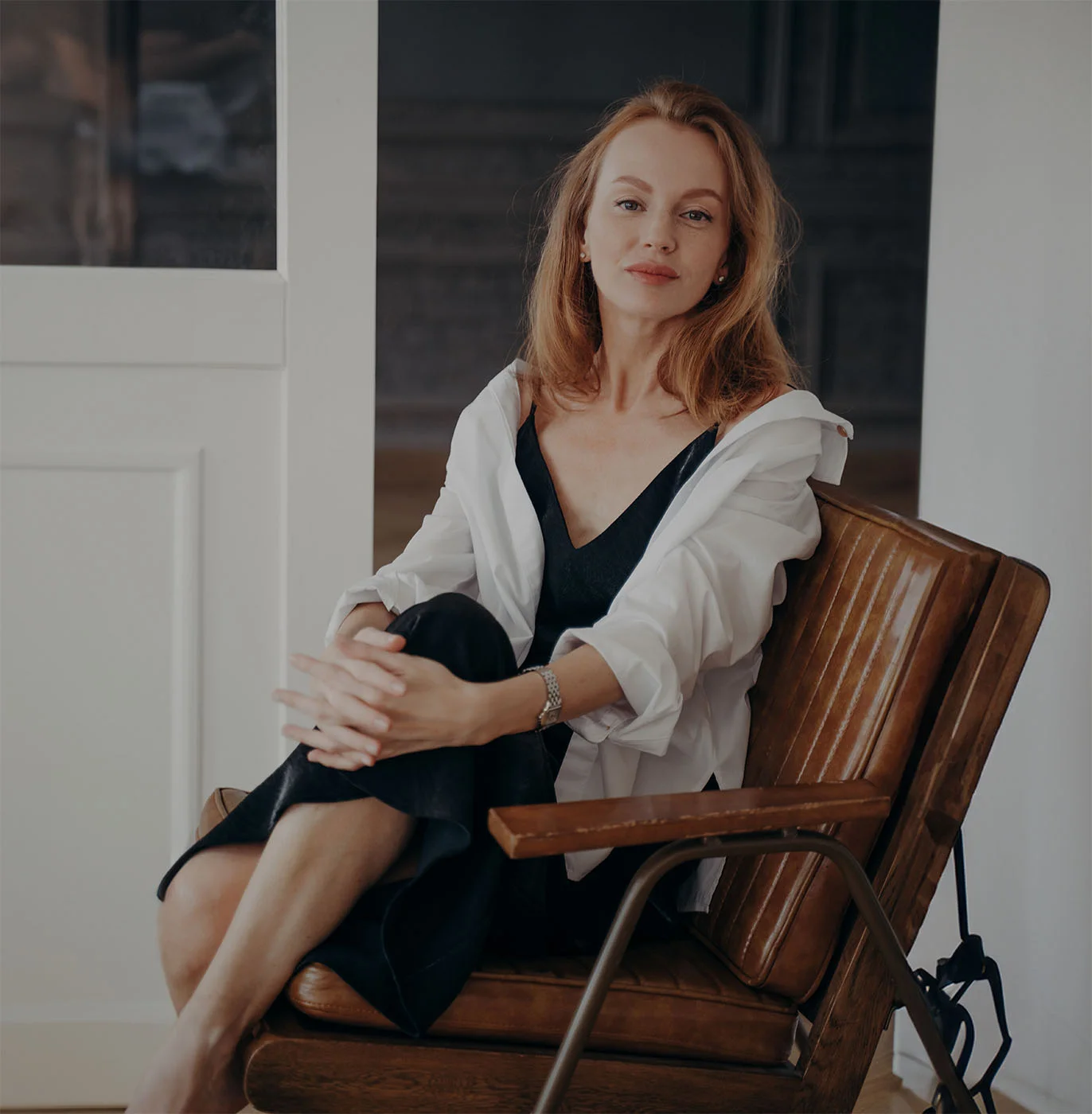 Portrait of a woman seated on a leather chair, slightly grinning at the camera. She has long, orange hair, and is wearing a black dress layered with a white long-sleeve shirt open over it. Her legs are crossed. - Womens Health in Webster, TX
