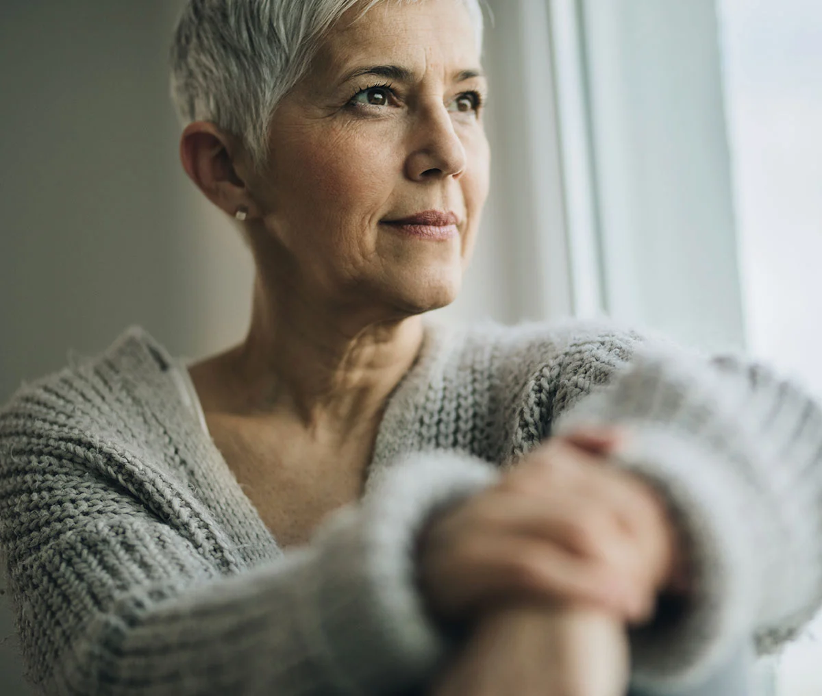 A close-up shot of a middle-aged woman with light makeup, gazing thoughtfully out a window. She has short hair and wears a cozy grey knit sweater. Her hands are clasped together within the frame, softly out of focus, adding a sense of warmth and introspection to the scene. - Kidney Cancer Treatment in Webster, TX