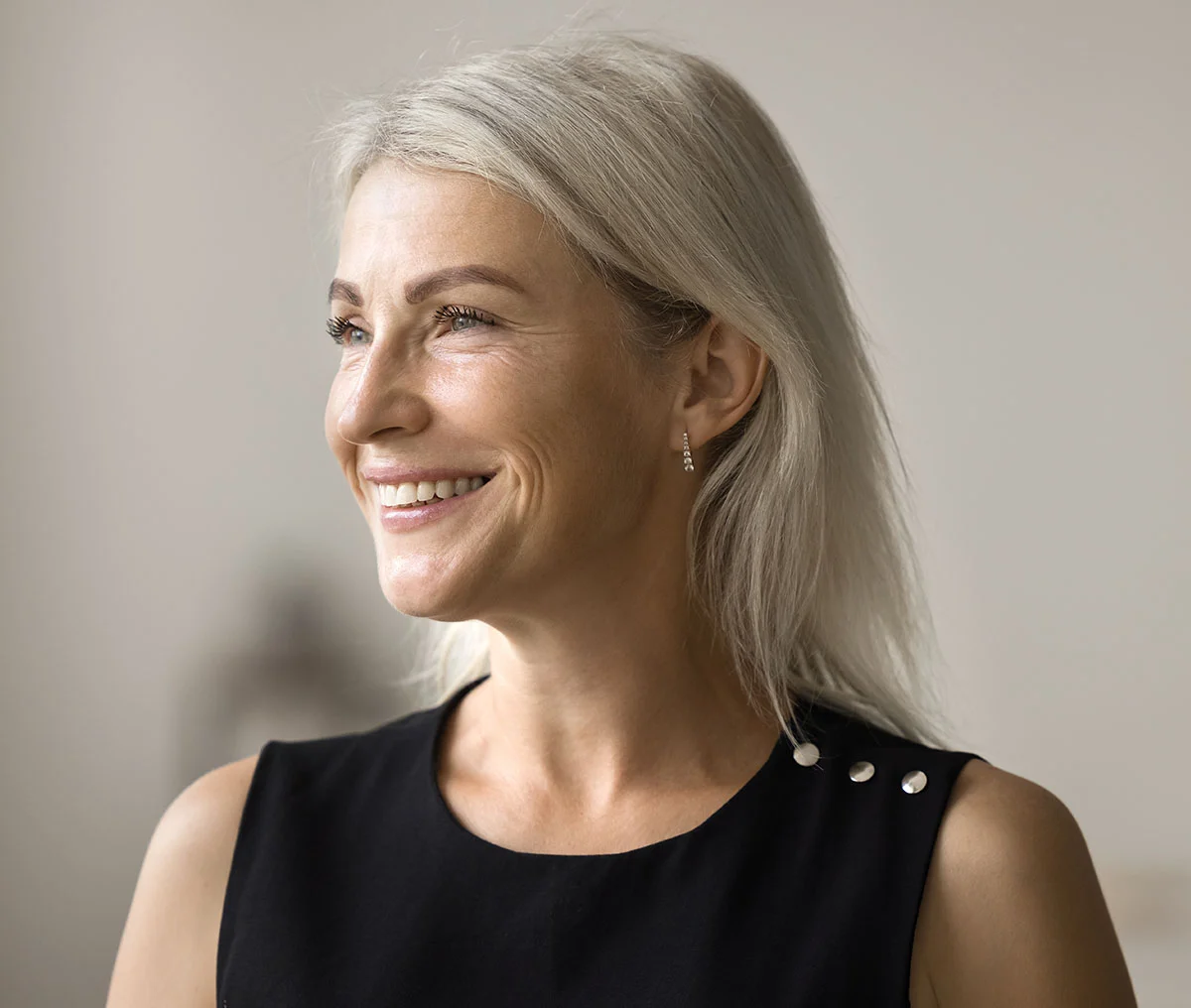 A close-up shot of a middle-aged woman smiling slightly, looking just to the side of the camera. She wears minimal makeup and a black sleeveless top, with her blonde hair styled softly. - Kidney Stone Disease (Women) Treatment in Webster, TX