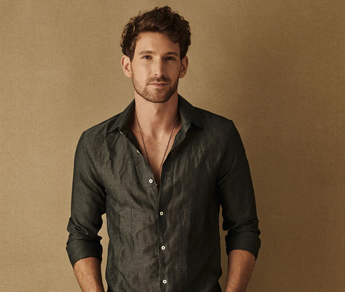 A young to middle-aged man stands in front of a light brown background. He has brown hair and a beard, wearing a grey long-sleeve button-down shirt with a few buttons open and the sleeves rolled up to his forearms. A chain is visible around his neck, adding to his casual, relaxed look. - Low Testosterone Treatment in Webster, TX