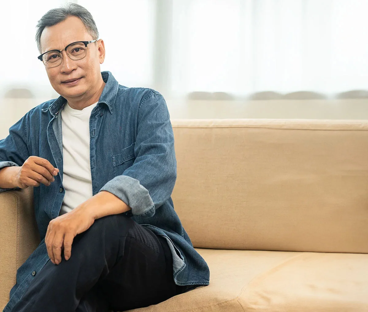 A middle-aged man sitting on a light brown couch, facing the camera with a slight smile. One arm rests on the arm of the chair, while the other is on his knee, crossed over his other leg. He is wearing black pants, a denim long-sleeve shirt rolled up to his forearms, a white undershirt, and glasses. - Prostate Biopsy in Webster, TX