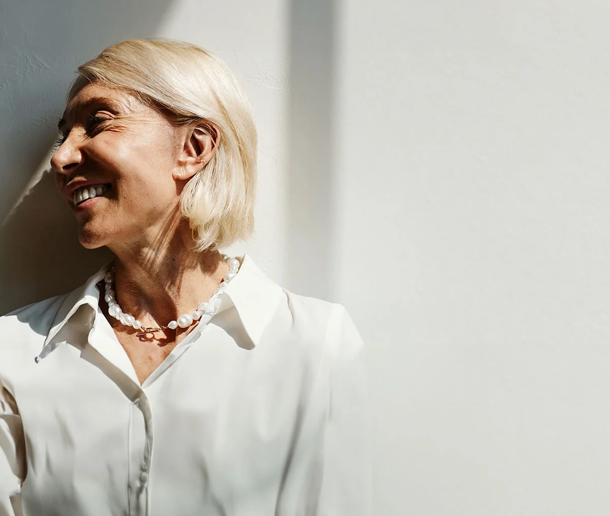 Close-up of a middle-aged woman wearing a white blouse and white pearls, smiling while looking away from the camera. She has little to no makeup and a blonde short bob hairstyle. - ProNox Nitrous Oxide Delivery System