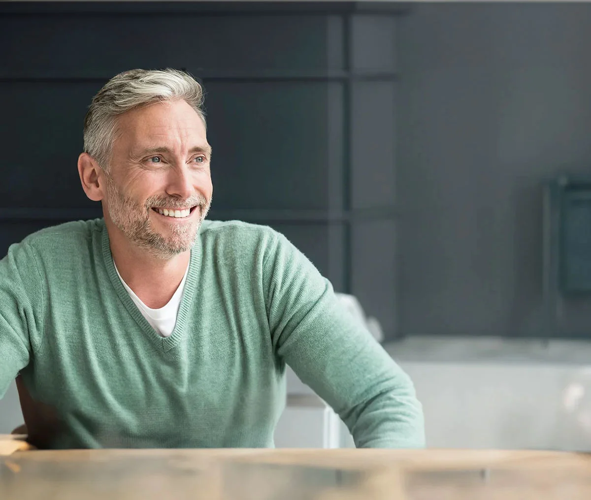 A man with light blonde hair and silver highlights in his beard and near his temples smiles broadly while looking to the left. Behind him is a sleek metal backdrop, adding a modern touch to the setting. He wears a green V-neck sweater layered over a white T-shirt, creating a relaxed and approachable look. - About Mens Health in Webster, TX