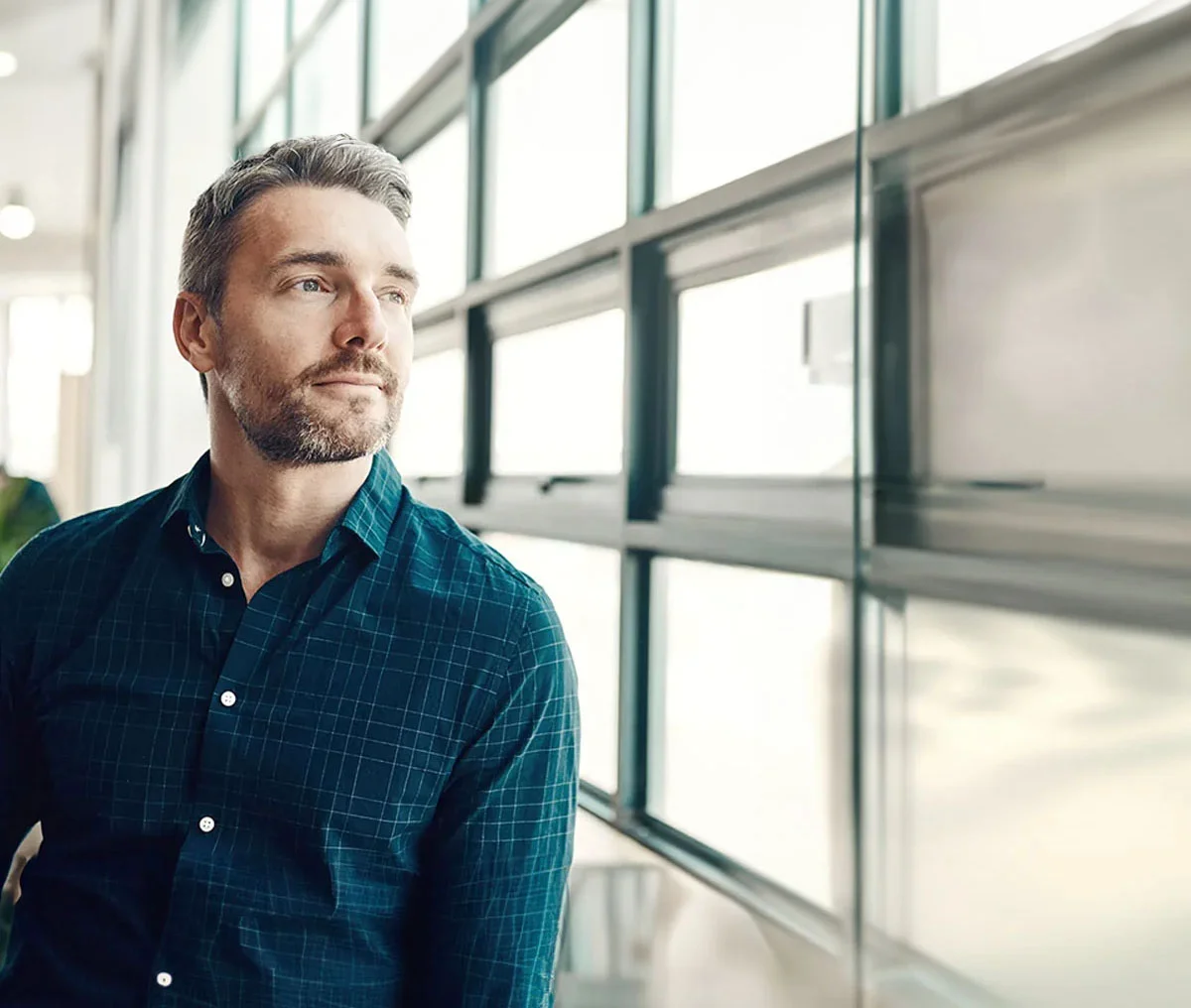 A middle-aged man with brown hair and a beard leans next to a wall full of windows. He is wearing a white dark drey-sleeve button-down shirt. With his arms crossed, he looks at the camera, conveying a thoughtful or confident expression. - Prostate Cancer Screening