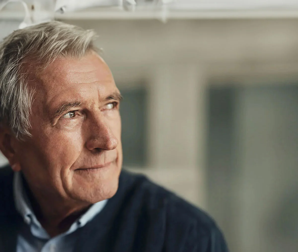 A close-up shot of an older white man with brown eyes, dressed in a light blue collared shirt beneath a navy sweater. He faces the camera but looks to the side, his expression neutral—neither smiling nor frowning. His grey hair adds to his mature appearance, and the background is softly blurred, keeping the focus on his thoughtful demeanor. - Elevated PSA Treatment in Webster, TX