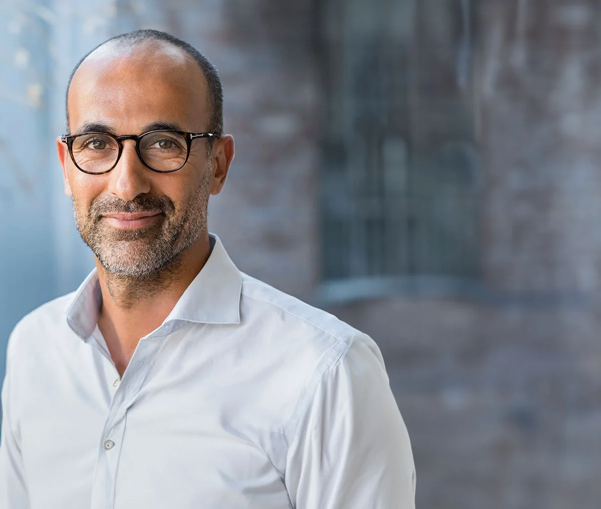 A close-up shot of a middle-aged man wearing a white collared button-down shirt and round-edged glasses. He smiles gently without showing his teeth, his scruff beard featuring brown and grey highlights. His buzzed hair is slightly receded, complementing his warm, approachable expression. - Erectile Dysfunction Treatment in Webster, TX