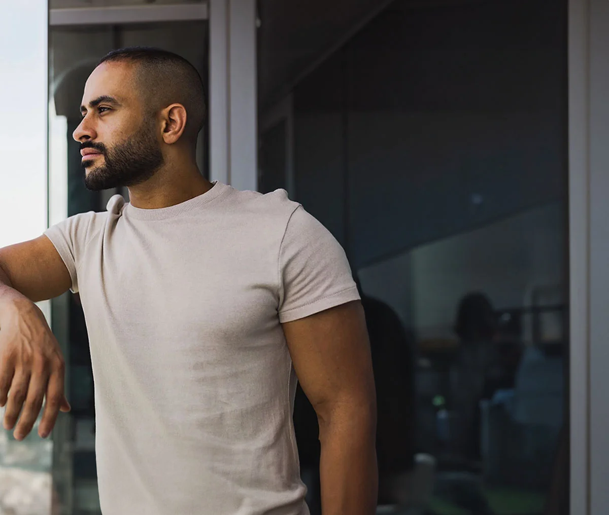 Semi-close-up shot of a Black man leaning slightly over a glass railing, looking out at the city. He has black hair and a beard, wearing a light beige shirt. - Male Infertility in Webster, TX