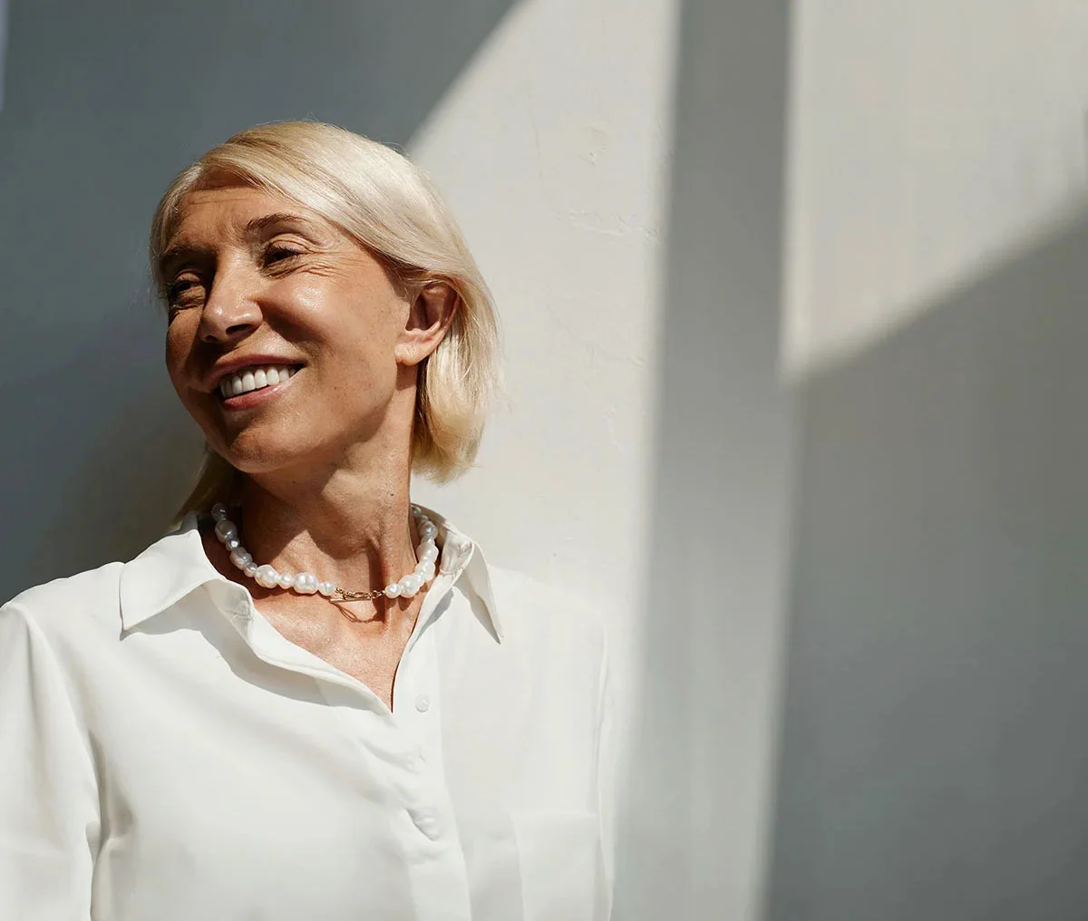 Shot of a middle-aged woman wearing a white blouse and white pearls, smiling while looking away from the camera. She has little to no makeup and a blonde short bob hairstyle. - Overactive Bladder