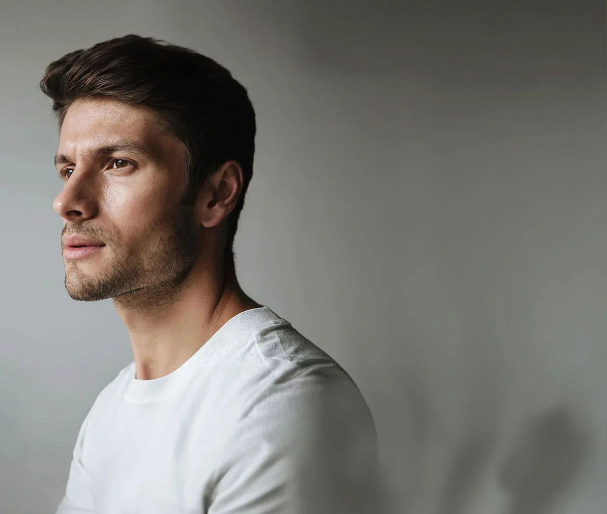 Shot of side profile of a middle-aged white man with brown hair and a thin beard. He is wearing a white t-shirt and looking slightly forward with a neutral expression. - Testicular Cancer