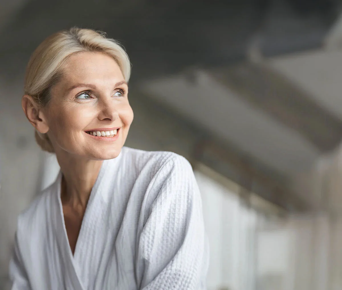 Photo of a middle aged women with blonde hair in a bun and white robe. Looking to the side and smiling. - Urinary Inontenence Women in Webster, TX