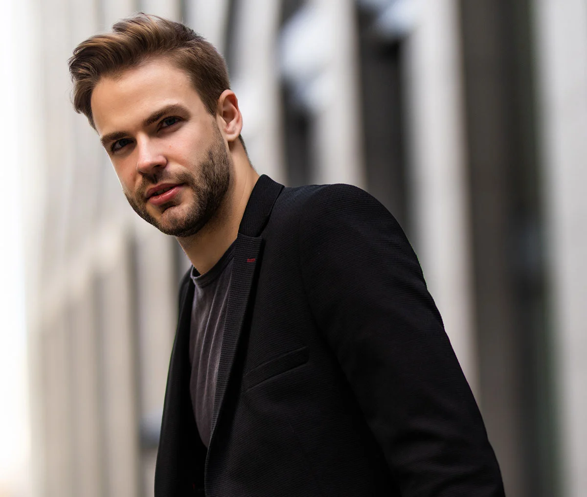 A young White male wearing a black blazer and dark grey t-shirt. He has light brown hair and a beard, looking directly into the camera. The background is blurred, but a building is visible. - Peyronies Disease Treatment in Webster, TX