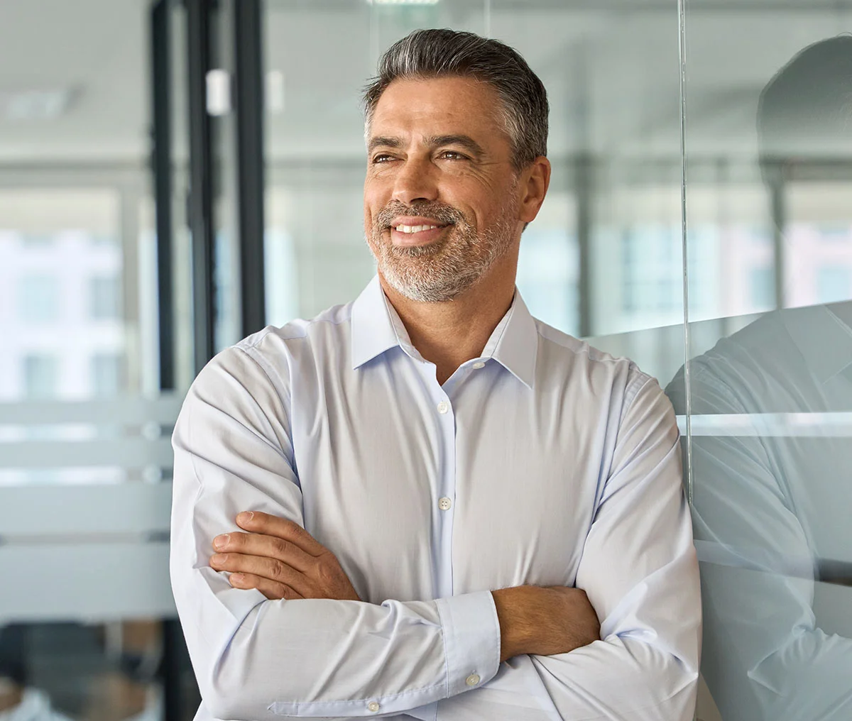 A middle-aged man with brown and white hair and a beard leans against a glass wall. He is wearing a white long-sleeve button-down shirt. With his arms crossed, he looks past the camera, conveying a thoughtful or confident expression. - Prostate Cancer