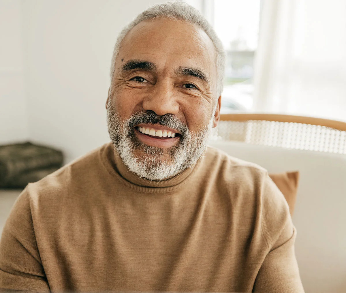 An older Black male genuinely smiling into the camera, wearing a brown sweater. He has white hair and a beard, with black highlights peeking through. - Ransurethral Resection of Prostate TURP