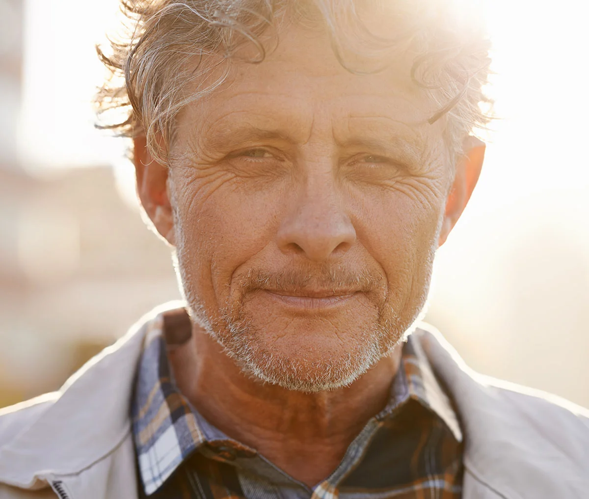 Close-up portrait of an older white man with grey hair and a thin beard. He is wearing a grey blazer over a multi-colored plaid shirt and looking slightly to the side with a neutral expression. - Stress Urinary Incontenence PSA