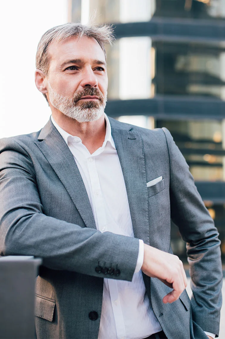 A middle aged man in a grey suit with a hand in the pocket and leaning against a railing, standing in front of a building. - Urilogical Cancer First Step in Webster, TX